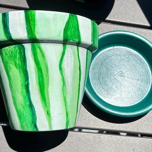 May include: A tapered plant pot featuring a hand-painted vertical striped pattern in varying shades of light and dark green, accompanied by a separate solid dark green circular saucer. The pot has a wide rim and a smooth, matte finish.