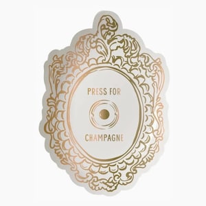 Pode incluir: Uma placa de parede branca com um design de moldura dourada ornamentada. O centro apresenta um botão circular dourado e o texto PRESS FOR CHAMPAGNE impresso em letras douradas. A placa tem uma borda recortada e um adorno decorativo superior.
