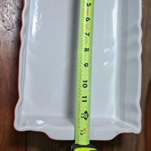 May include: A rectangular white ceramic baking dish with scalloped edges. A yellow measuring tape is placed vertically inside the dish, showing a length of approximately 13 inches from the top inner edge to the bottom inner edge of the basin.