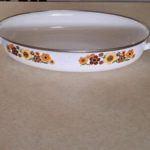 May include: An oval-shaped white enamel roasting pan featuring a speckled finish and a decorative floral pattern in shades of brown, orange, and yellow. The pan includes two side handles and a thin metallic rim around the top edge of the vessel.