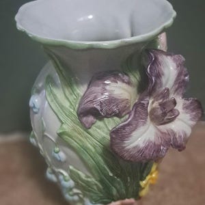 May include: A ceramic vase with a scalloped rim, featuring three-dimensional sculpted floral details including a large purple iris, green leaves, and small light blue flowers against a white background with a glossy finish and textured surface.