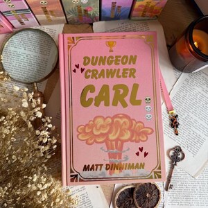 Op de afbeelding: Een roze hardcover boek getiteld Dungeon Crawler Carl door Matt Dinniman, met een gouden trofee-icoon, een illustratie van een paddenstoelwolk en drie kleine schedeliconen op de voorkant, omlijst door een gouden rand met kleine rode hartjes.