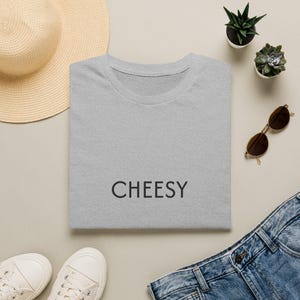 Puede incluir: Una camiseta de manga corta en color gris jaspeado con la palabra CHEESY impresa en letras mayúsculas negras en la parte delantera. La prenda se muestra extendida, con cuello redondo y un diseño sencillo y limpio para uso diario.