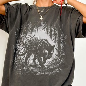 May include: A charcoal gray oversized t-shirt featuring a black and white graphic print of a panther prowling through a dense forest. The shirt has short sleeves, a crew neckline, and a faded, washed-out appearance. The word PEPPER is printed below.