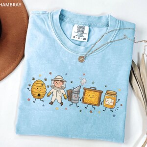 以下が含まれることがあります： 明るいブルーのTシャツで、蜂の巣、養蜂家、燻煙器、巣枠、蜂蜜の瓶が描かれたグラフィックプリントが施されています。すべて笑顔のキャラクターで、Comfort Colorsのラベルが付いており、小さな星の模様が散りばめられています。