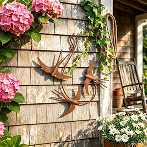 May include: A wall-mounted metal sculpture featuring three stylized swallows in a rusted brown finish, arranged in a circular formation with a decorative scroll hook at the top, designed for outdoor display on a wooden shingle exterior wall.