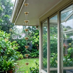 Op de afbeelding: Een goudkleurige metalen regenketting met een reeks verbonden trechtervormige bekers die verticaal aan een dakrand hangen. Tijdens een regenbui stroomt het water door de open bekers naar beneden, wat een functioneel en decoratief afvoersysteem vormt.