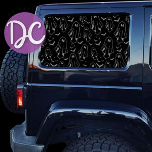 May include: A black vehicle rear side window decal featuring a repeating pattern of white outlined ghosts and small bats. The design covers the glass surface, and a circular purple logo with the white text DC is positioned in the upper left corner.