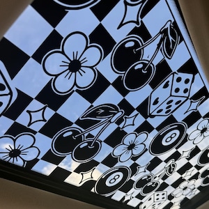 May include: A translucent car sunroof decal featuring a black and white checkered pattern overlaid with black graphic illustrations of cherries, flowers, eight balls, dice, and four-pointed stars, allowing natural light to filter through the design.
