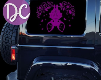 Puede incluir: Un adhesivo de vinilo color magenta vibrante aplicado en la ventana trasera de un vehículo negro. El diseño muestra un murciélago estilizado colgado boca abajo de una rama, rodeado de flores, pequeñas estrellas y siluetas de murciélagos.