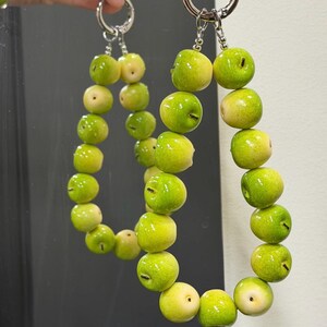 Könnte beinhalten: Ein Paar Schlüsselanhänger bestehend aus einer Reihe kleiner, glänzender, hellgrüner und gelber apfelförmiger Perlen. Jeder Schlüsselanhänger ist an einem silberfarbenen Metallring befestigt und bildet eine dekorative Schlaufe aus Fruchtanhängern.