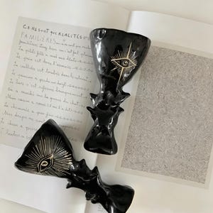 Könnte beinhalten: Zwei schwarze Keramikkelche mit unregelmäßigen, organischen Formen und glänzenden Oberflächen. Jeder Kelch ist mit einem handgemalten goldenen Auge mit strahlenförmigen Linien verziert. Die Gefäße haben eine skulpturale Silhouette und einen breiten Rand.