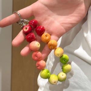 Könnte beinhalten: Ein Schlüsselanhänger mit einer Reihe kleiner, glatter, kugelförmiger apfelförmiger Perlen, die in einem Farbverlauf angeordnet sind. Die Perlen gehen von Dunkelrot oben zu Orange, Gelb und schließlich Hellgrün unten über und sind an einem Metallring befestigt.
