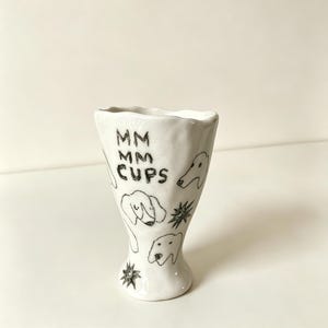May include: A white ceramic cup with an irregular, wavy rim and a flared base. The exterior features hand-drawn black ink illustrations of dog faces, star-like shapes, and the text MM MM CUPS written in a casual, block-letter style font.
