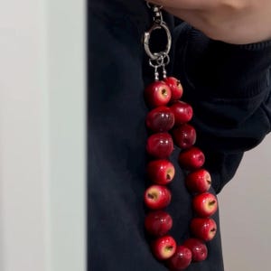 Könnte beinhalten: Ein Schlüsselanhänger mit einer Reihe kleiner, glänzender, apfelförmiger roter Perlen, die aneinandergereiht sind. Die Äpfel sind an einem silberfarbenen Metallring mit Verschluss befestigt und bilden ein hängendes Accessoire in leuchtendem Rot.