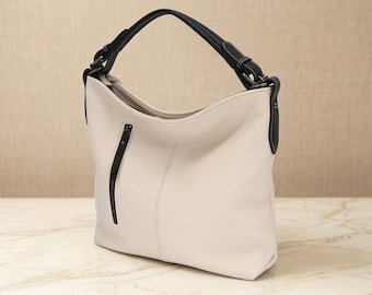 May include: A hobo-style shoulder bag in off-white with a dark navy blue handle and trim. The bag features a vertical zippered pocket on the front, a single top handle attached with dark hardware, and visible stitching along the seams and handle.