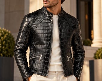 Puede incluir: Una chaqueta de cuero negro con textura de piel de cocodrilo, que presenta un cuello alzado, una cremallera central plateada y dos bolsillos laterales con cremallera. La chaqueta se lleva sobre una camisa blanca y pantalones claros.