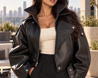 May include: A black leather jacket with a wide collar, silver-tone zipper, and snap button closures on the cuffs and hem. The jacket features long, loose sleeves and a belted waist detail. It is worn over a plain cream-colored top and black trousers.