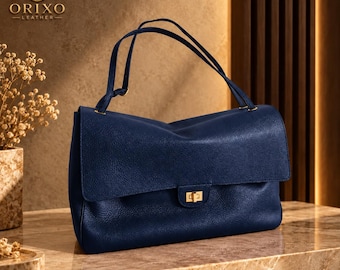 May include: A navy blue handbag with a textured surface, featuring a fold-over flap, a gold-toned rectangular turn-lock closure, and two thin shoulder straps. The logo ORIXO LEATHER is visible in the upper left corner of the image.