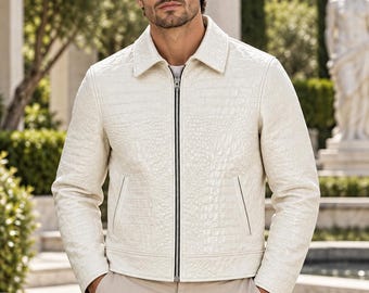 May include: An off-white leather jacket featuring a textured crocodile-embossed pattern, a classic collar, and a full-length silver-tone front zipper. The garment includes two side pockets and long sleeves, with the ORIXO LEATHER logo visible.