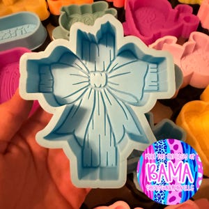 May include: A light blue plastic cookie cutter shaped like a decorative bow with detailed embossed lines. The cutter features a central knot and flowing ribbon ends. A circular sticker with the text Find me on Etsy at BAMA SCENTSANDMORELLC is visible.