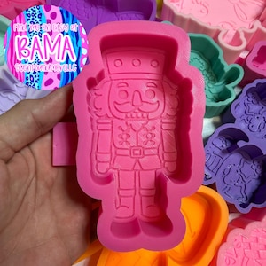 May include: A bright pink silicone mold featuring a detailed nutcracker design with a tall hat, uniform jacket, and boots. The mold has a raised outline of the nutcracker figure, providing a clear impression for crafting projects.