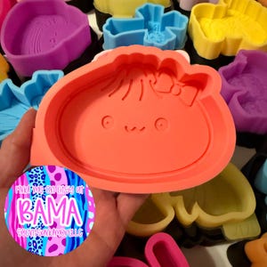 May include: A bright orange silicone mold featuring a rounded character with a small bow, two circular eyes, and a curved mouth. The mold is surrounded by various other colorful silicone molds in shades of blue, yellow, and purple, with a circular sticker displaying the text Find me on Etsy at BAMA SCENTSANDMORELLC.