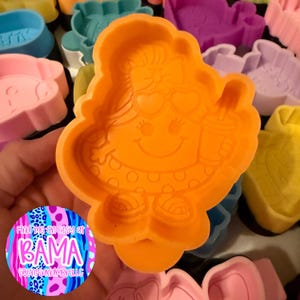 May include: An orange plastic cookie cutter featuring a cartoon character with a bow, heart-shaped sunglasses, and a drink. The character wears a polka-dot skirt and sandals. The design is embossed into the center of the orange plastic frame.