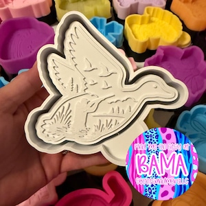 May include: A white plastic cookie cutter shaped like a duck in flight with a hunter figure in the foreground. The design features embossed details of wings, reeds, and a landscape. A circular sticker with the text Find me on Etsy at BAMA SCENTSANDMORELLC is attached to the lower right corner.