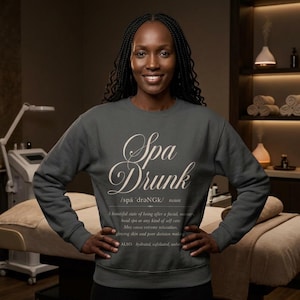 May include: A dark gray crewneck sweatshirt featuring the white text Spa Drunk, a phonetic pronunciation, and a dictionary-style definition describing a state of relaxation after a facial, massage, or self-care session with long sleeves.