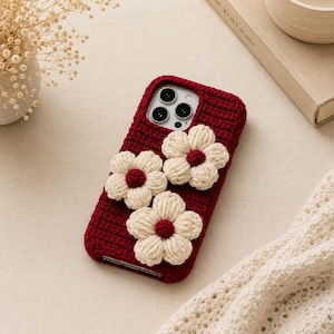 May include: A dark red crochet phone case featuring three dimensional cream-colored flowers with dark red centers. The case has a textured, ribbed knit pattern and a cutout for the camera lens array on the upper left side of the rectangular cover.
