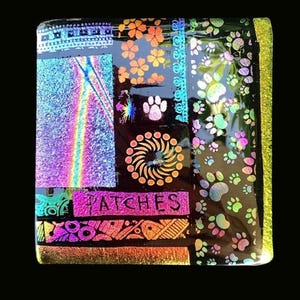May include: A square glass tile featuring a black background with iridescent, multi-colored patterns including floral shapes, paw prints, a circular sun-like design, and a rectangular section with the word PATCHES printed in black lettering.
