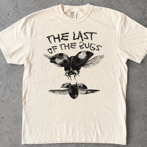 Könnte beinhalten: Ein cremefarbenes Kurzarm-T-Shirt mit einem schwarzen Grafikdruck von zwei fliegenden Marienkäfern. Über den Insekten steht der Text THE LAST OF THE BUGS in einer abgenutzten, handgezeichneten Schriftart. Das Shirt hat einen Rundhalsausschnitt.