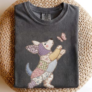 以下が含まれることがあります： チャコールグレーの半袖Tシャツ。花柄やチェック柄の布地をパッチワークした犬のグラフィックが描かれており、小さなピンクの蝶に手を伸ばしています。首元の内側のラベルにはComfort Colorsの文字が記されています。