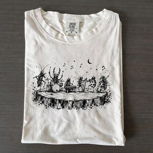 Może przedstawiać: Kremowy t-shirt z czarną ilustracją przedstawiającą owady grające na instrumentach muzycznych na pniu drzewa. Grafika zawiera świerszcza, chrząszcza, pszczołę, biedronkę i osę wraz z nutami, gwiazdami oraz półksiężycem nad nimi.