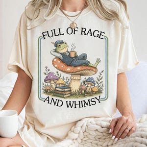 Könnte beinhalten: Ein cremefarbenes T-Shirt mit einer Grafik eines Frosches in blauer Weste und Mütze, der auf einem großen Pilz liegt und eine Tasse hält. Unter dem Frosch befinden sich gestapelte Bücher und kleinere Pilze. Der Text lautet FULL OF RAGE AND WHIMSY.