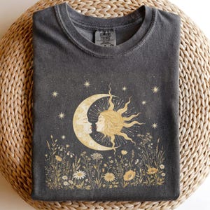 Könnte beinhalten: Ein anthrazitfarbenes T-Shirt mit Rundhalsausschnitt und einem Grafikdruck, der einen goldenen Halbmond und eine Sonne mit menschlichen Gesichtern zeigt, umgeben von Sternen und Wildblumen. Das Innenetikett trägt die Aufschrift Comfort Colors.