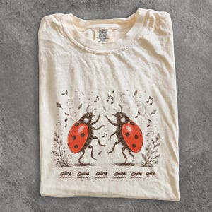 May include: A cream-coloured t-shirt featuring a graphic print of two ladybirds dancing. The ladybirds have bright red wings with black spots. Surrounding them are brown illustrations of musical notes, leaves, and a row of ants at the bottom.