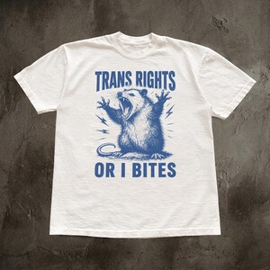 May include: A white short-sleeved t-shirt featuring a blue graphic of a snarling opossum with its claws raised. Above the opossum, the text reads TRANS RIGHTS, and below the opossum, the text reads OR I BITES, with small lightning bolt accents.