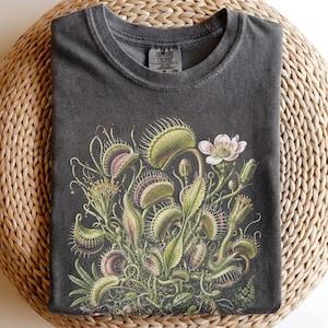 Puede incluir: Una camiseta de cuello redondo gris carbón con una ilustración botánica detallada de varias plantas atrapamoscas con trampas verdes y rosas, tallos largos y una sola flor rosa pálido. La etiqueta interior muestra el texto Comfort Colors.