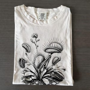 Peut inclure: Un t-shirt à manches courtes blanc cassé avec une illustration détaillée à l'encre noire d'une plante Dionaea muscipula. Le t-shirt présente un col rond et une étiquette Comfort Colors imprimée à l'intérieur avec la taille M visible.