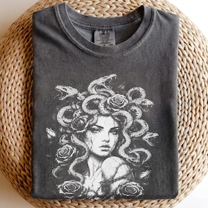 Puede incluir: Una camiseta de cuello redondo gris carbón con un estampado gráfico blanco de una figura con serpientes por cabello, rodeada de rosas. La etiqueta interior del cuello muestra el texto Comfort Colors, indicando la marca y la talla.