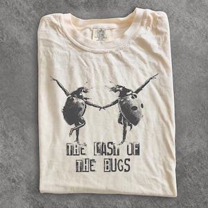 Może przedstawiać: Kremowy t-shirt z czarną grafiką przedstawiającą dwie biedronki stojące na tylnych odnóżach i trzymające się za ręce. Poniżej owadów znajduje się napis THE LAST OF THE BUGS wykonany w postarzanym, blokowym kroju pisma.