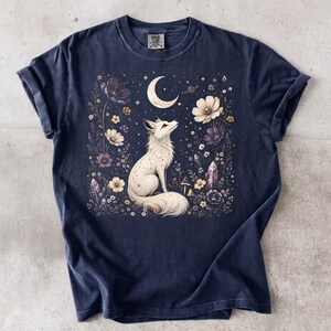 Puede incluir: Una camiseta azul marino de manga corta con un gráfico central de un zorro blanco sentado entre flores, cristales y una luna creciente. El diseño incluye pequeñas estrellas y elementos celestiales. La prenda tiene cuello redondo y etiqueta.