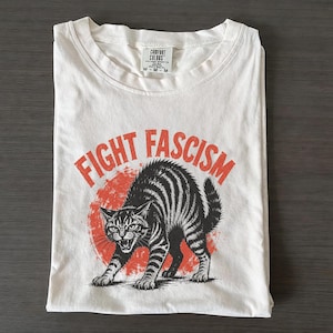 Könnte beinhalten: Ein cremefarbenes T-Shirt mit einer Grafik einer fauchenden, schwarz gestreiften Katze mit gekrümmtem Rücken vor einem verwaschenen roten Kreis. Der Text "FIGHT FASCISM" ist in einer fetten, roten, gebogenen Schrift über dem Katzenmotiv gedruckt.