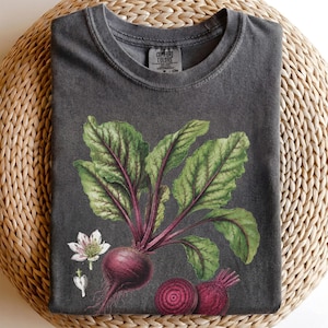 May include: A charcoal grey crew neck t-shirt featuring a botanical illustration of a whole beetroot with large green leaves, a small white flower, and two sliced beetroot halves. The inner neck label displays the Comfort Colors brand name.
