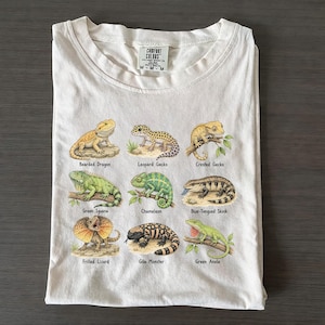 Könnte beinhalten: Ein cremefarbenes T-Shirt mit einem Grafikdruck von neun verschiedenen Echsen in einem Raster. Die Abbildungen zeigen eine Bartagame, einen Leopardgecko, einen Kronengecko, einen Grünen Leguan, ein Chamäleon, einen Blauzungenskink, eine Kragenechse, eine Gila-Krustenechse und einen Karolina-Anolis, jeweils mit dem Namen darunter gedruckt.