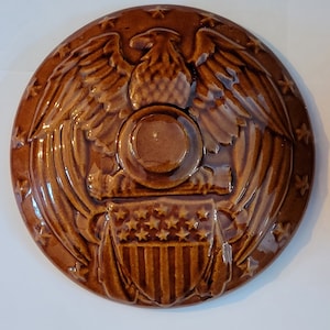 Peut inclure: Un couvercle en céramique brun avec un motif en relief d'un aigle aux ailes déployées, un bouclier avec des étoiles et des rayures, et un cercle avec des étoiles autour.