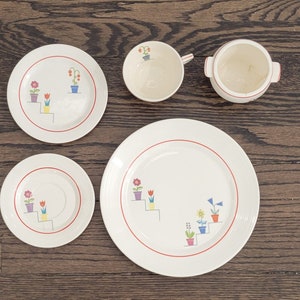 May include: A vintage set of white ceramic dishes with a red rim and a floral design featuring potted plants and flowers. The set includes a large dinner plate, a smaller salad plate, a saucer, a teacup, and a creamer.