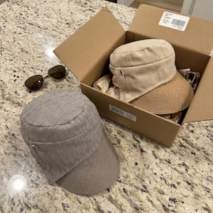 May include: Two cadet-style caps with a structured crown and a woven brim. One cap is light beige and sits inside a cardboard shipping box, while the other is heather gray and rests outside. Both feature a silver-tone safety pin on the side.